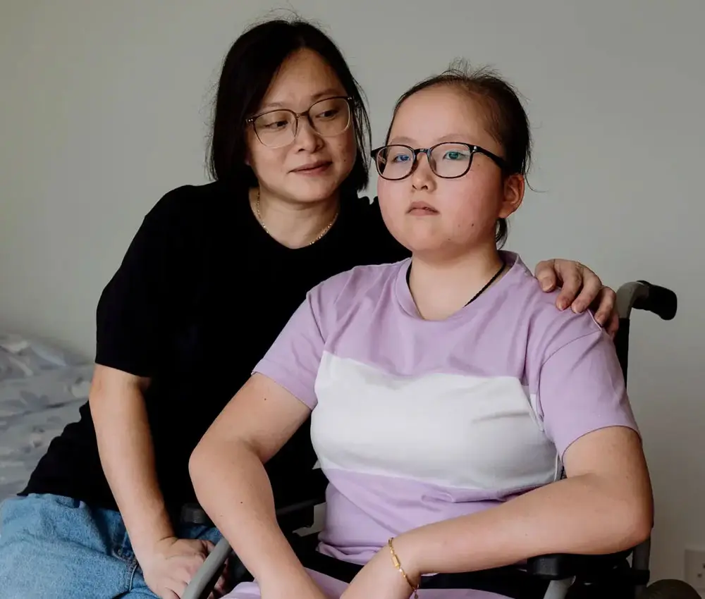 A child in a wheelchair sits with her mother in a bedroom.