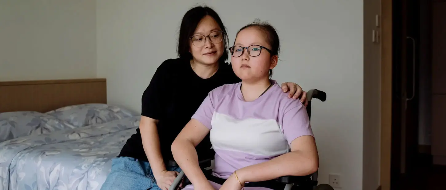 A child in a wheelchair sits with her mother in a bedroom.